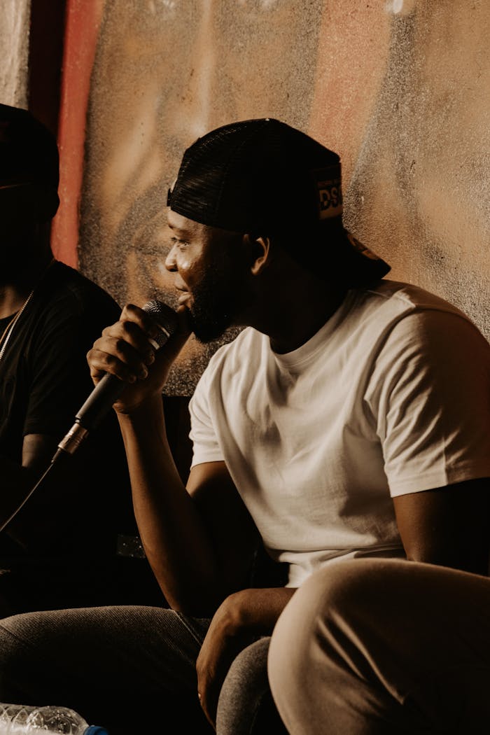 who-we-are Black man in cap holding microphone in casual setting, singing or rapping.