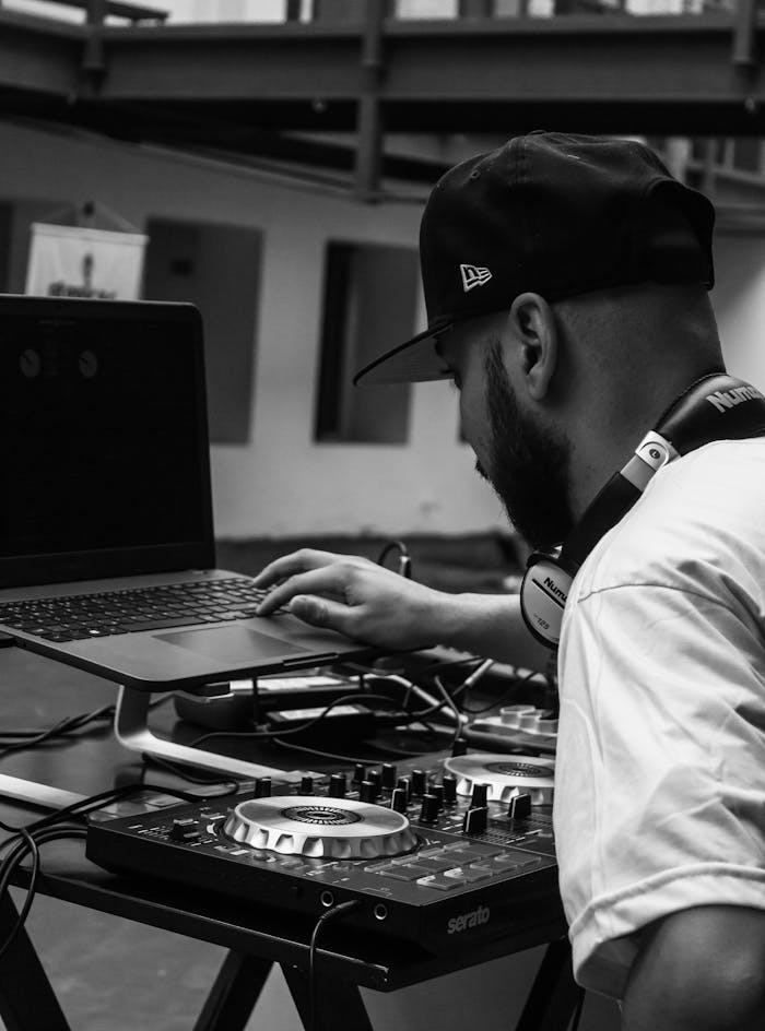 Black and white photo of a DJ using a laptop and mixer indoors, creating music.