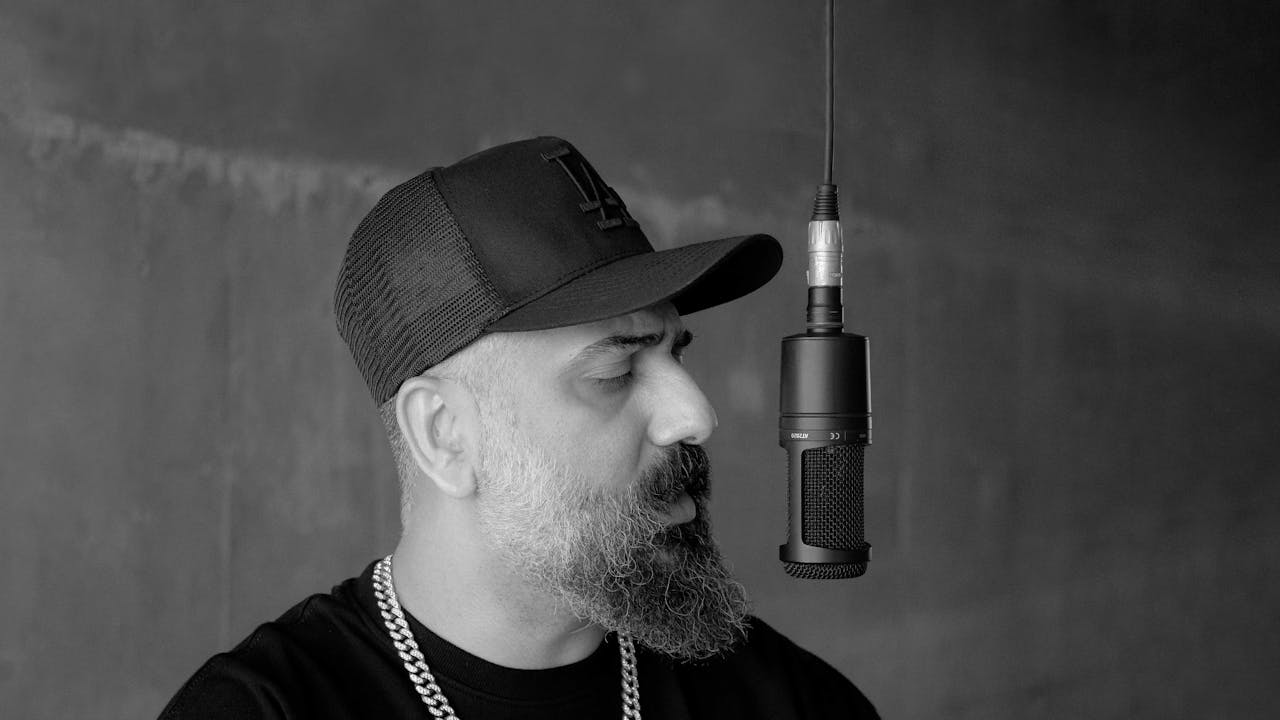 services-01 Black and white portrait of a bearded rapper wearing a cap in front of a microphone.