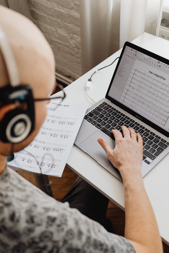 services-03 Adult musician composing music on a laptop using score sheets and headphones.